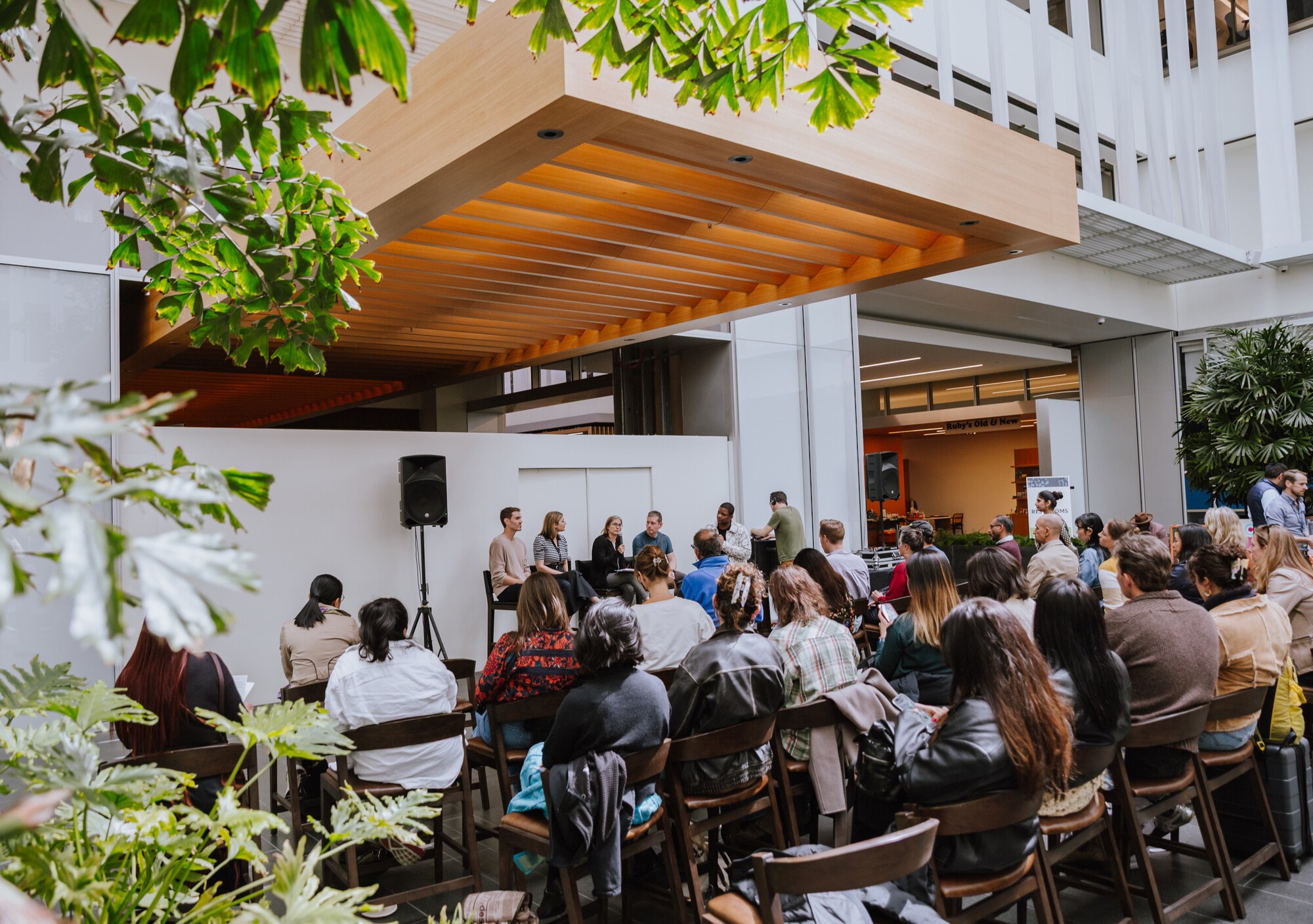 A panel discussion under the wood canopy in the One Market Plaza atrium, the audience seated among the plants